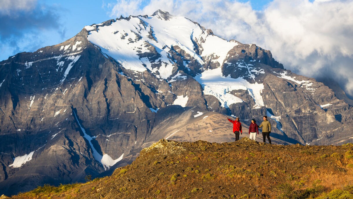 Three hikers stand on a rocky ridge pointing toward the dramatic snow-capped peaks of Torres del Paine National Park in Chilean Patagonia.