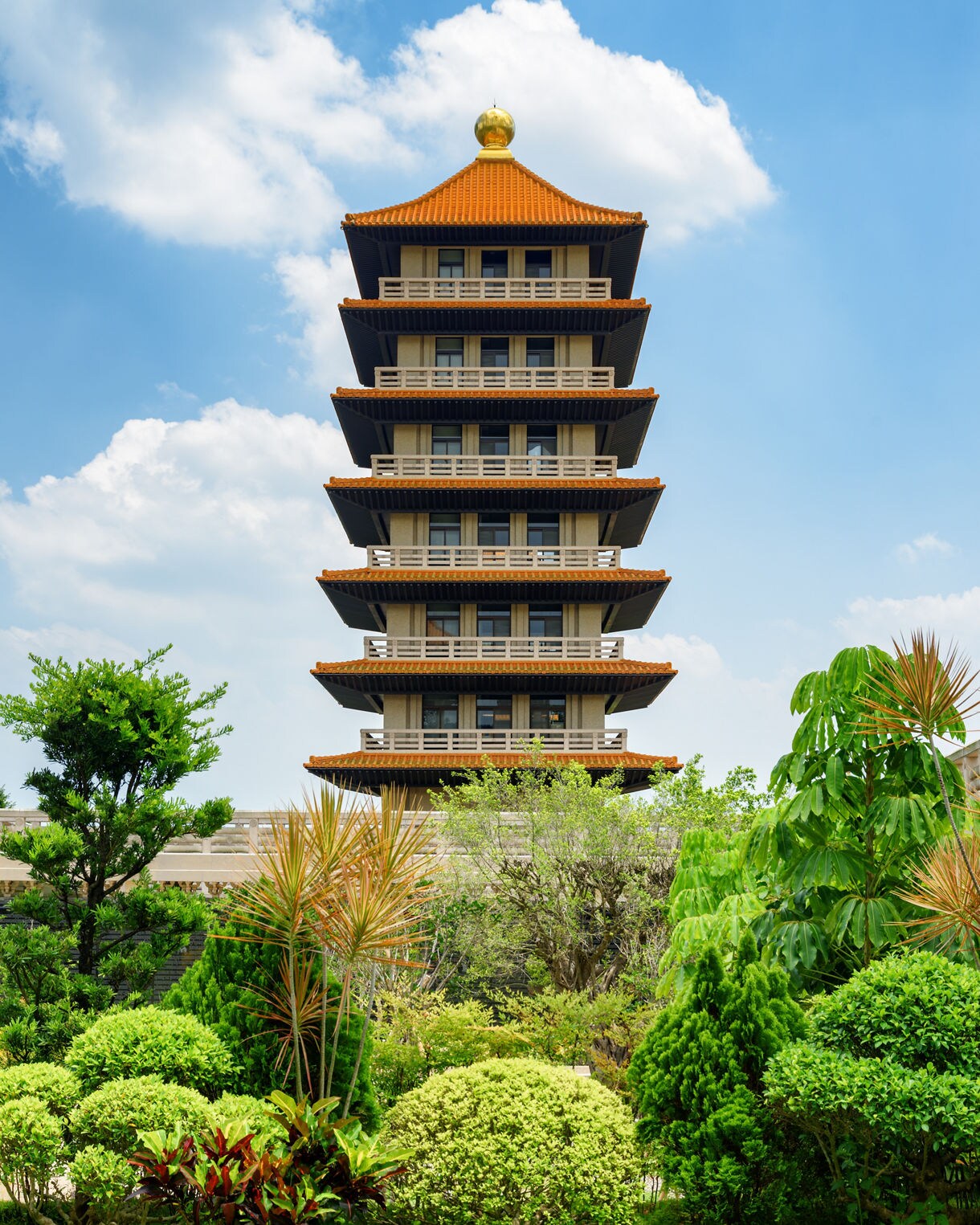 A multi-tiered pagoda with an orange-tiled roof and golden finial at Fo Guang Shan Monastery in Kaohsiung, Taiwan, surrounded by lush green gardens under a bright sky.