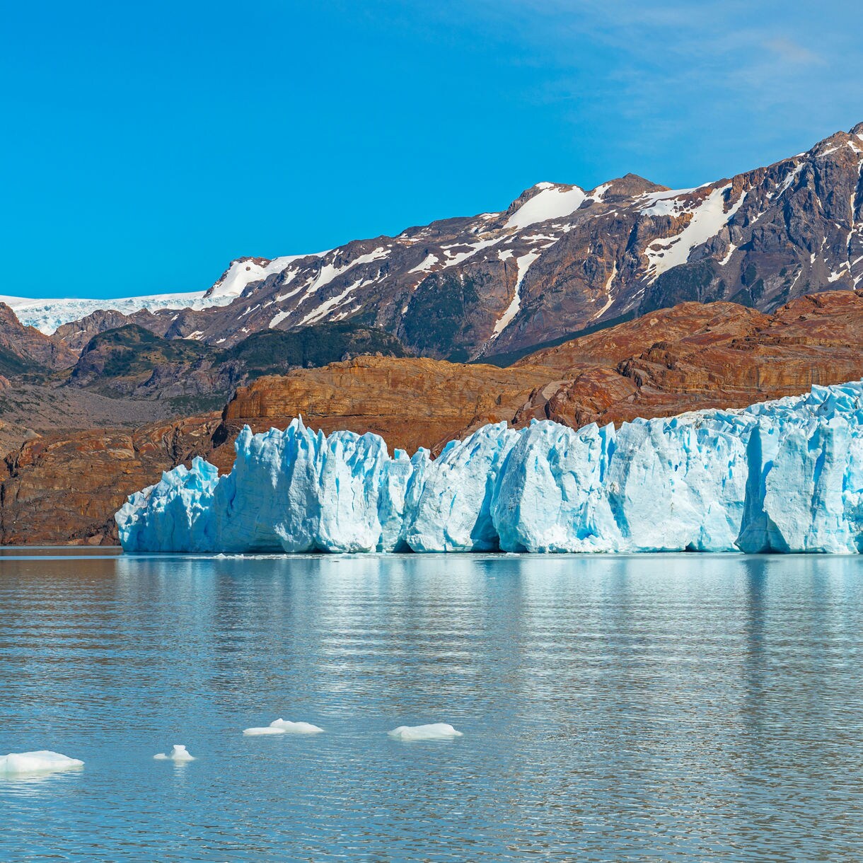 Expansive view of Grey Glacier in Patagonia, showing bright blue ice cliffs meeting calm water with rocky mountains and patches of snow in the background.