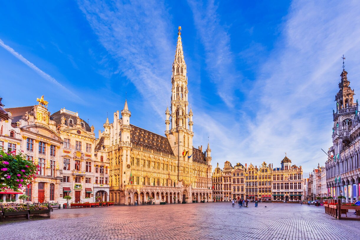 Evening view of Brussels’ Grand Place with the illuminated Town Hall tower, ornate guildhalls and cobblestone square under a colorful sunset sky.