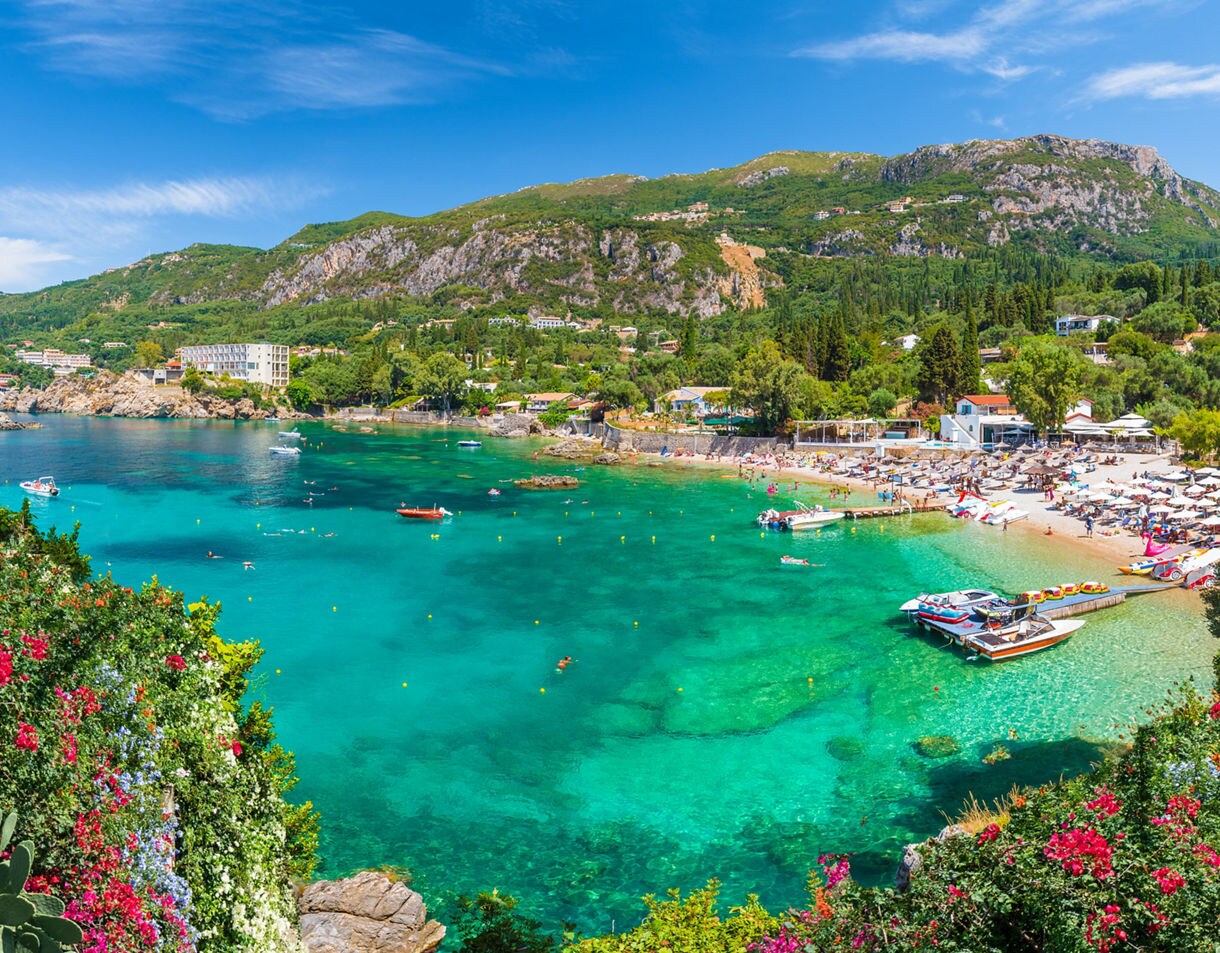 Vibrant view of Paleokastritsa Bay in Corfu, Greece, with clear turquoise water, sandy beach lined with umbrellas, boats anchored offshore and lush green hills in the background.
