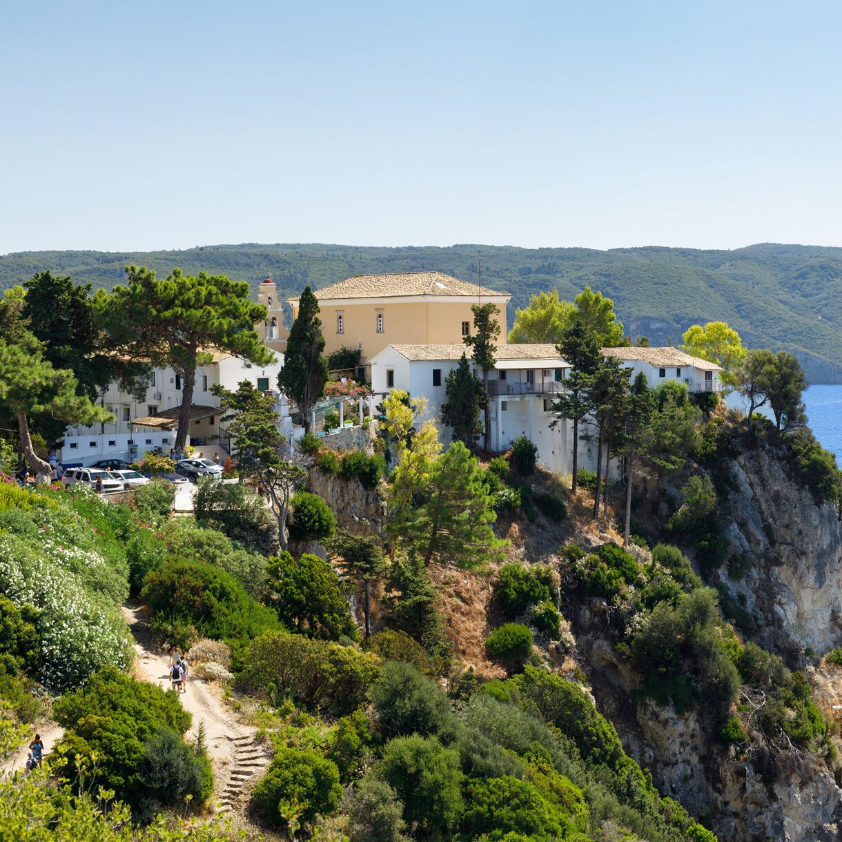Paleokastritsa Monastery on a cliffside in Corfu, surrounded by lush greenery, overlooking the deep blue Ionian Sea and nearby islands.