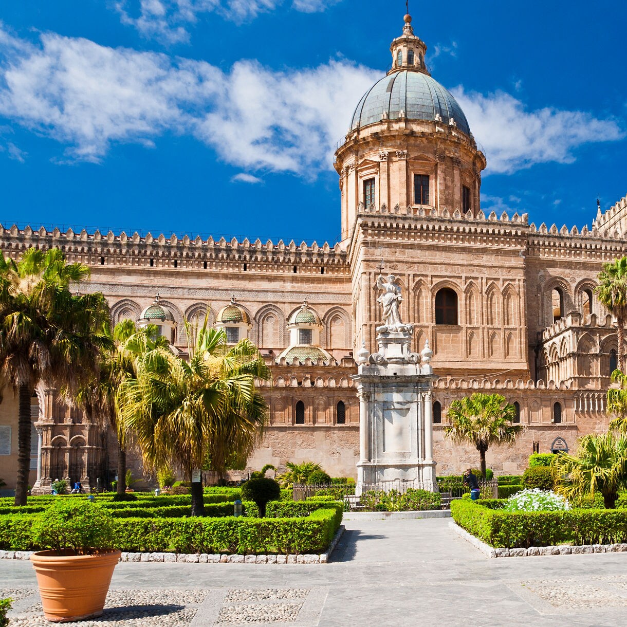 Exterior view of Palermo Cathedral with its large dome, ornate stone facade and surrounding palm-filled garden under a bright blue sky.