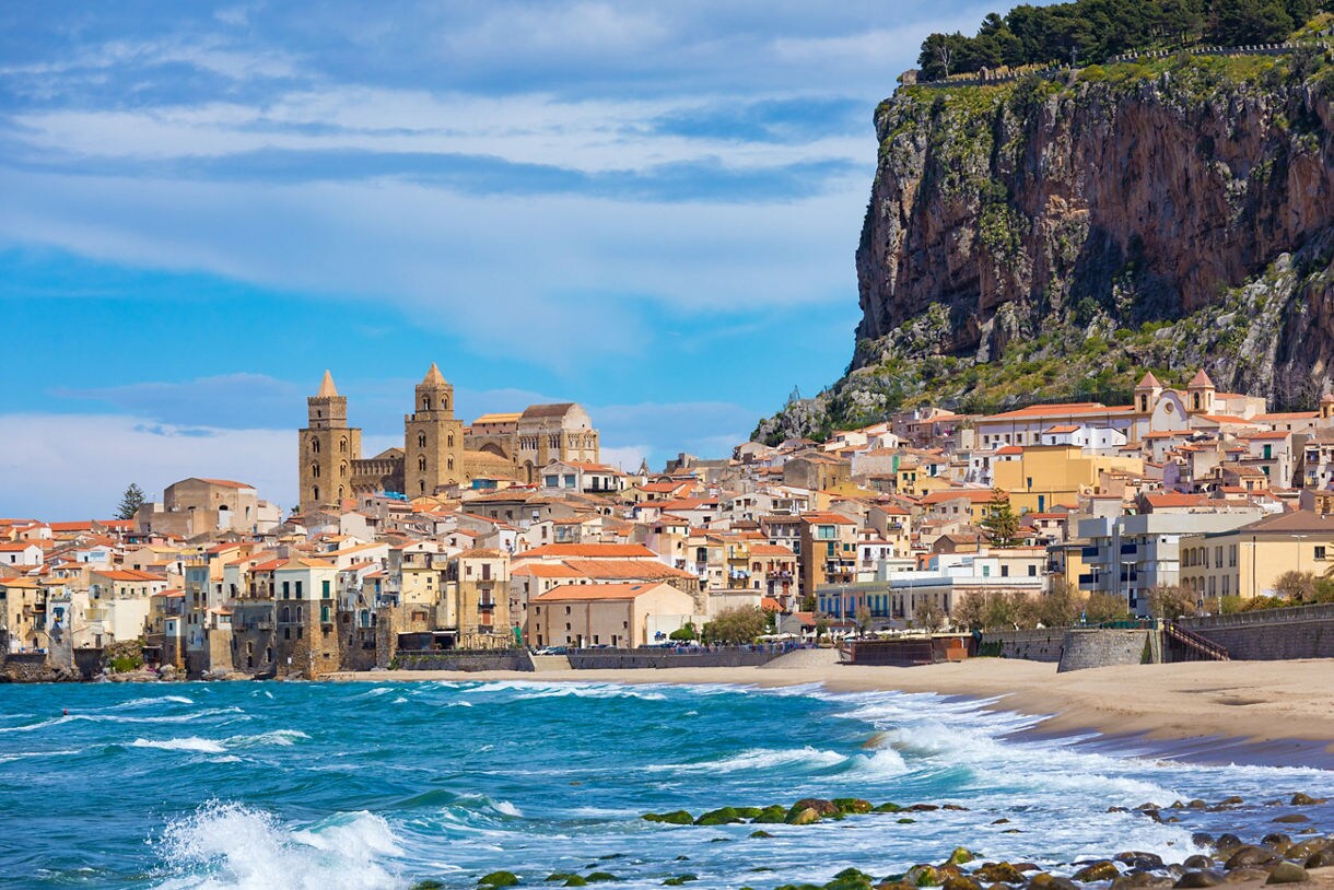 Coastal town of Cefalù with orange rooftops, a large medieval cathedral and waves crashing along the sandy beach below a rocky cliff.