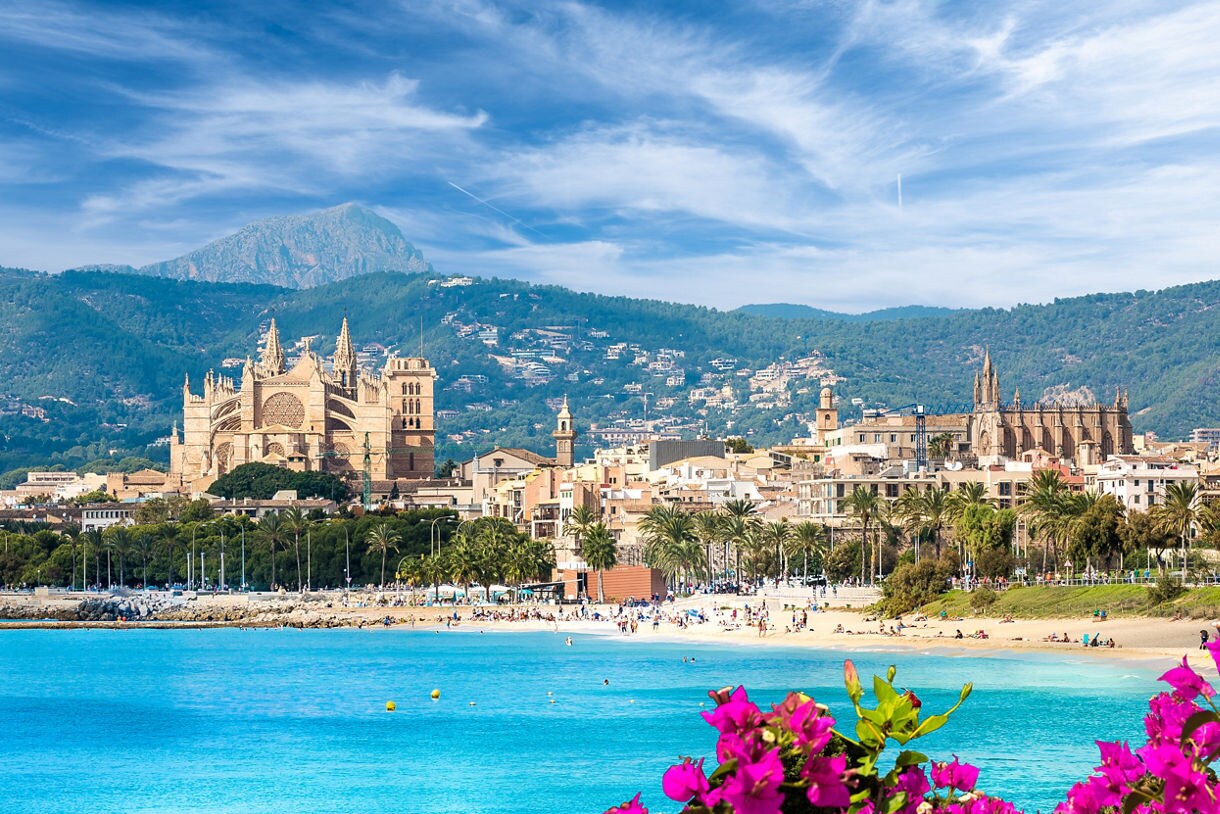 View of Palma de Mallorca, Spain, featuring La Seu Cathedral near the beach with turquoise water, palm trees and green mountains in the background.