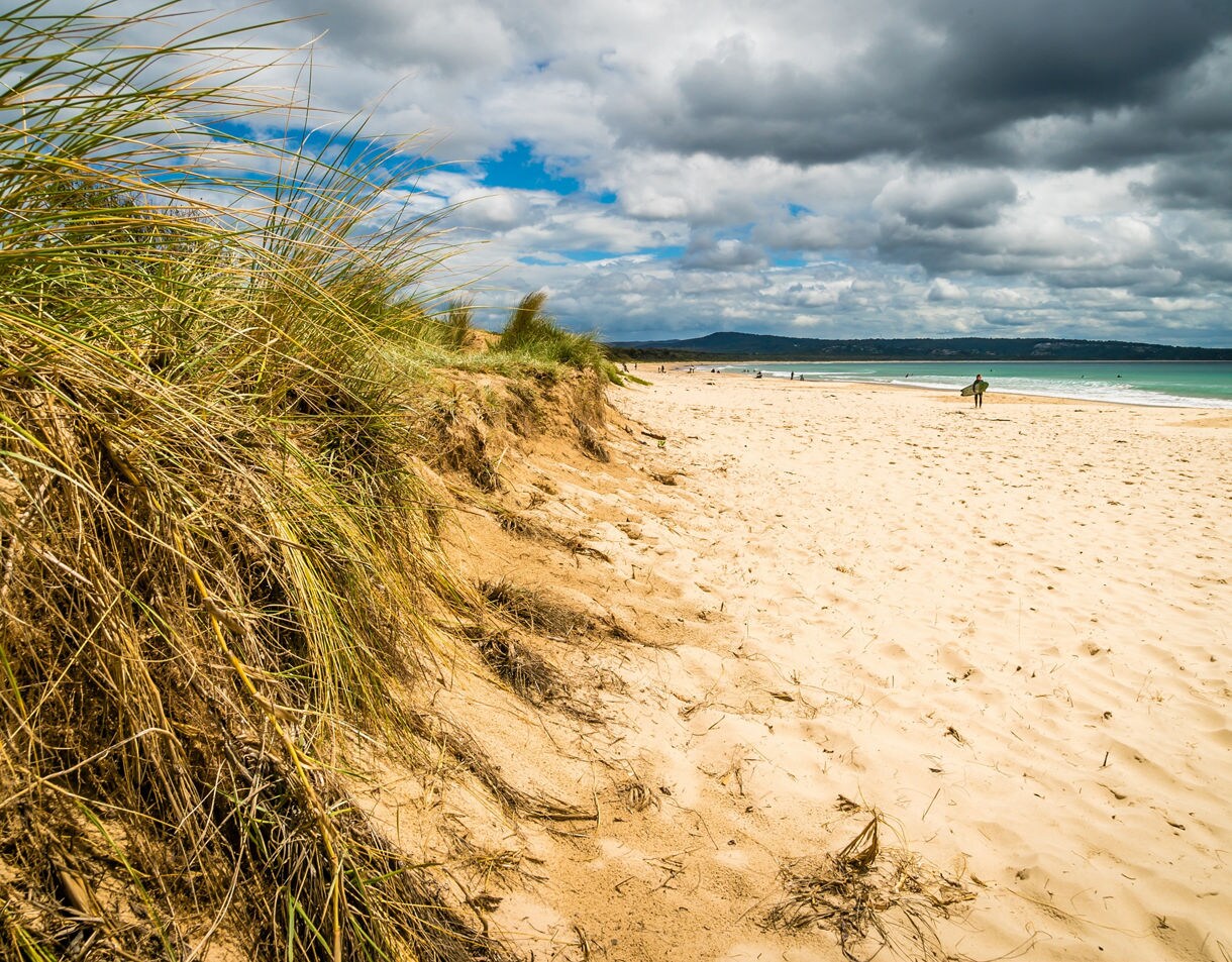 Golden sand beach with patches of dune grass in the foreground, gentle waves and a distant figure walking near the shoreline under partly cloudy skies.