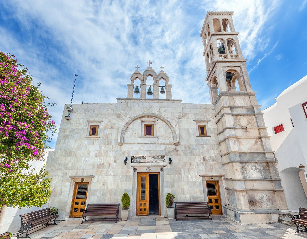 White marble facade of Panagia Tourliani Monastery in Mykonos, featuring arched windows, three bells with crosses and a detailed bell tower beside blooming flowers.