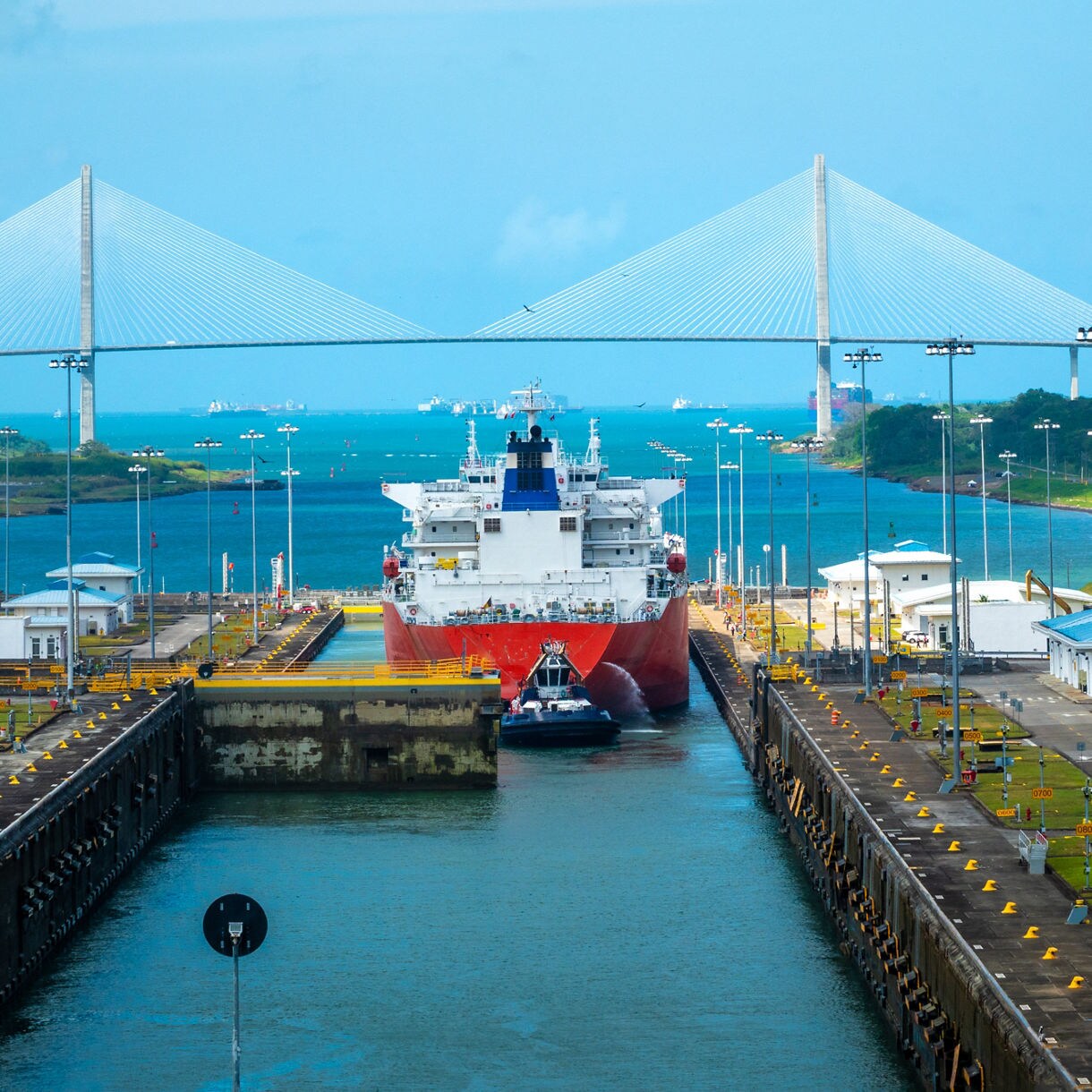 A large cargo ship navigates through the locks of the Panama Canal, assisted by tugboats. The setting features a vibrant blue sky, modern infrastructure, and the iconic cable-stayed Centennial Bridge in the background. The scene highlights maritime engineering and global trade routes.