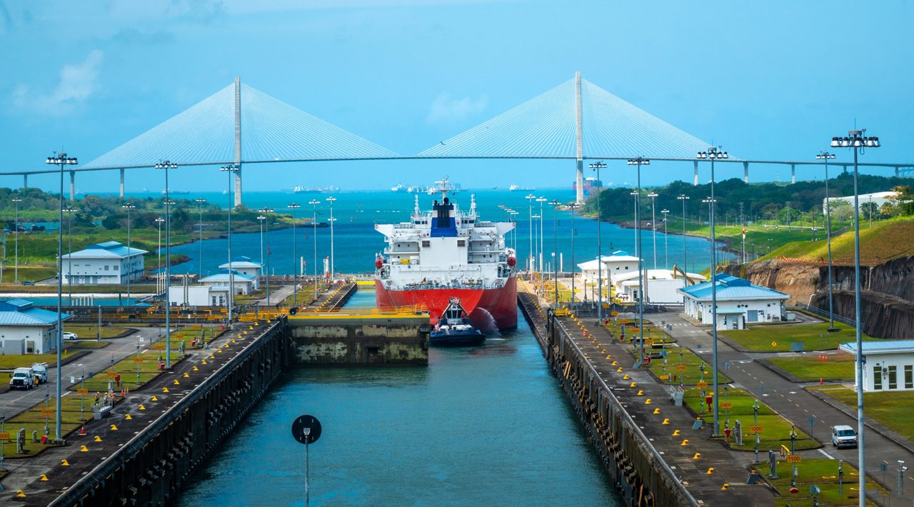 Large cargo ship passing through the Panama Canal locks with a suspension bridge and turquoise waters in the background.