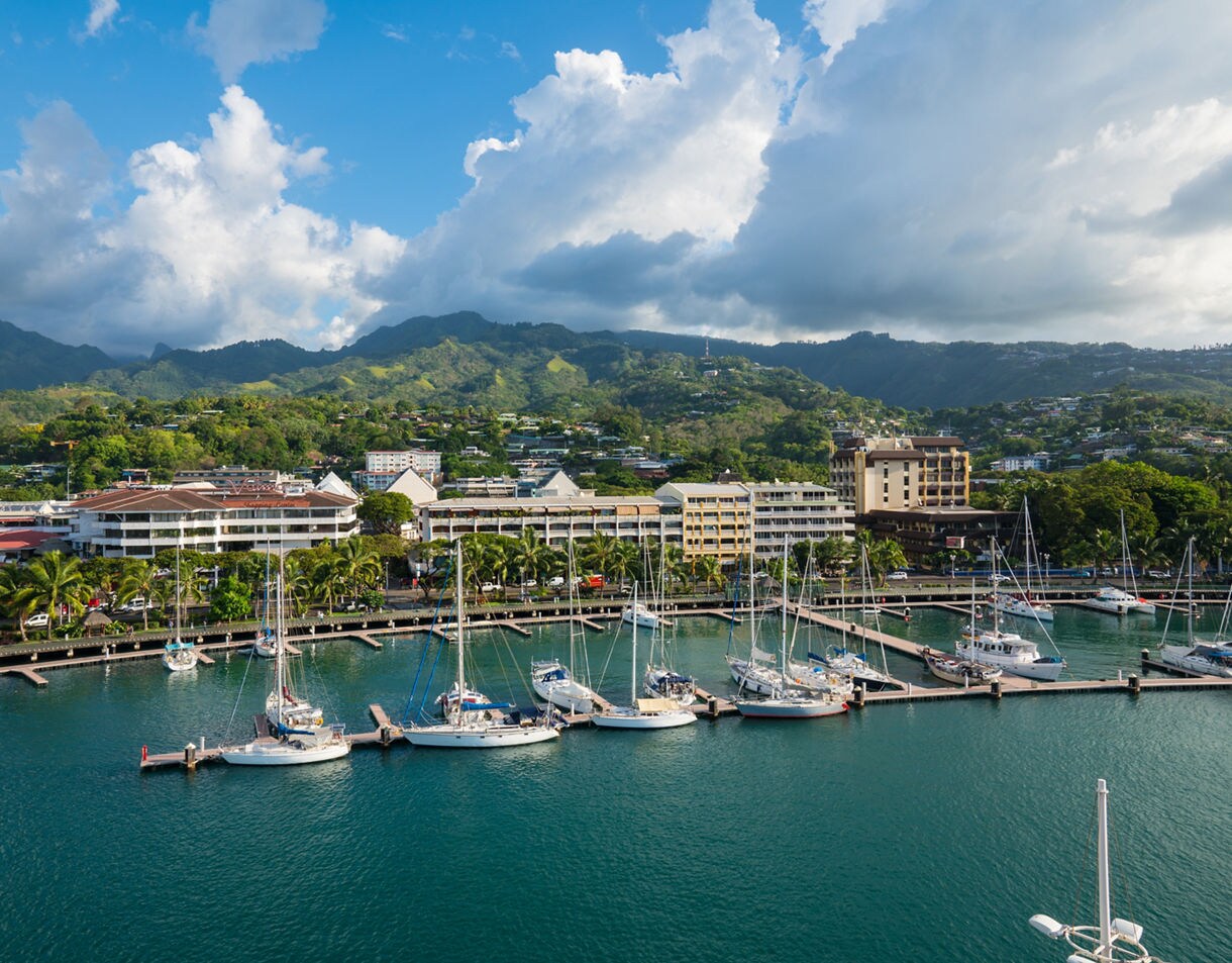 Marina in Papeete with rows of sailboats docked beside palm-lined waterfront buildings backed by lush green mountains under bright clouds.