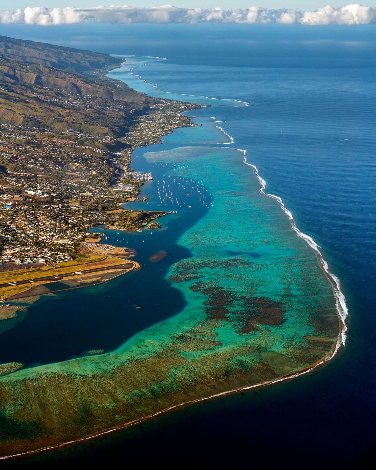 Aerial view of Tahiti’s coastline with vibrant turquoise reefs, a long white surf break, deep blue ocean and coastal towns backed by rugged green mountains.
