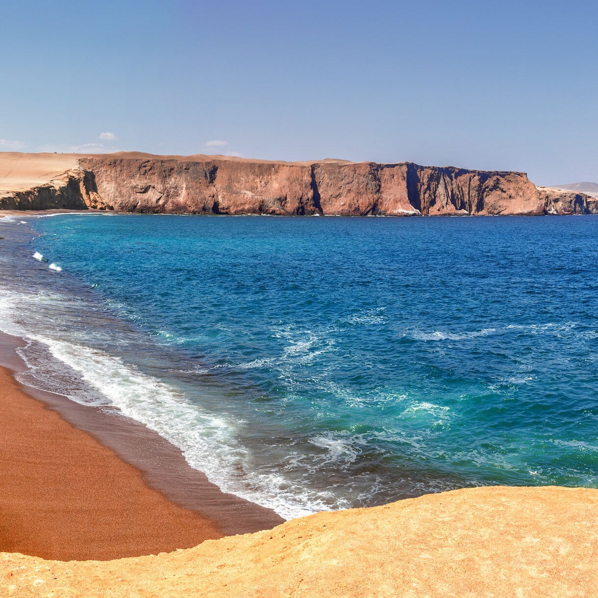 Coastal scene in Paracas National Reserve with red sand beach, rugged cliffs and bright blue ocean under a clear sky.