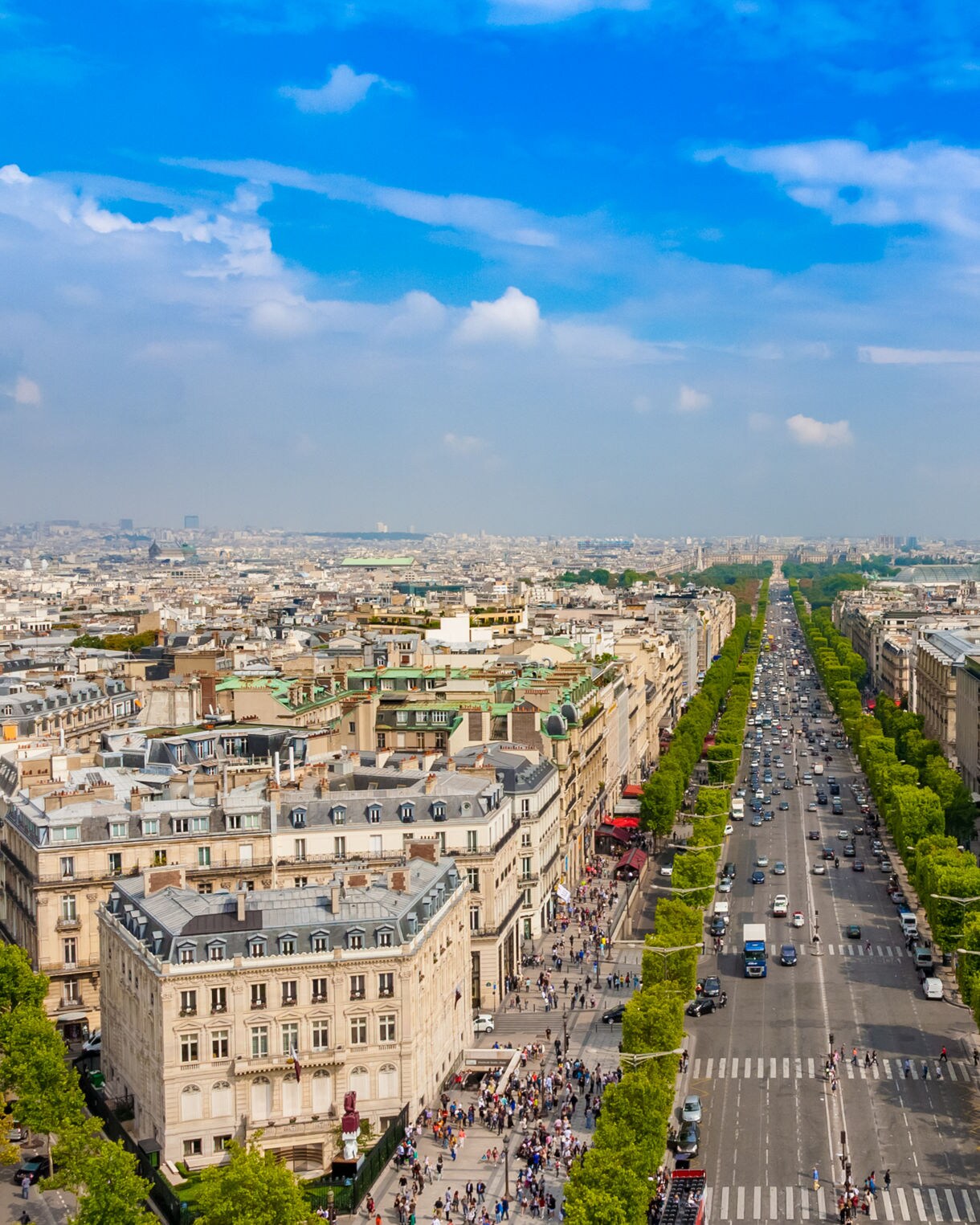 Aerial view of the Champs-Élysées in Paris, showing wide tree-lined boulevards filled with cars and pedestrians, stretching into the distance.
