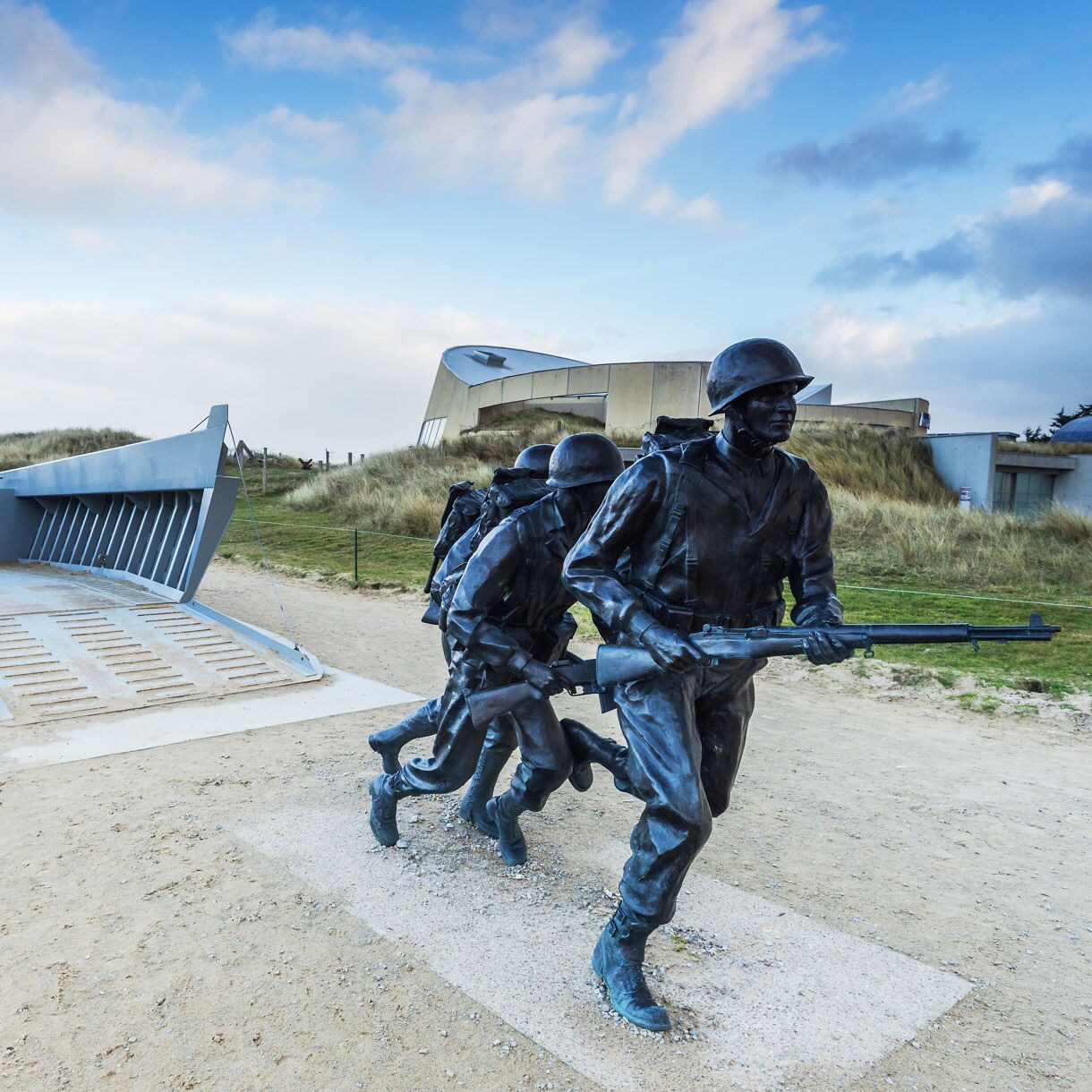 Bronze statues of World War II soldiers advancing beside a landing craft on Normandy Beach.