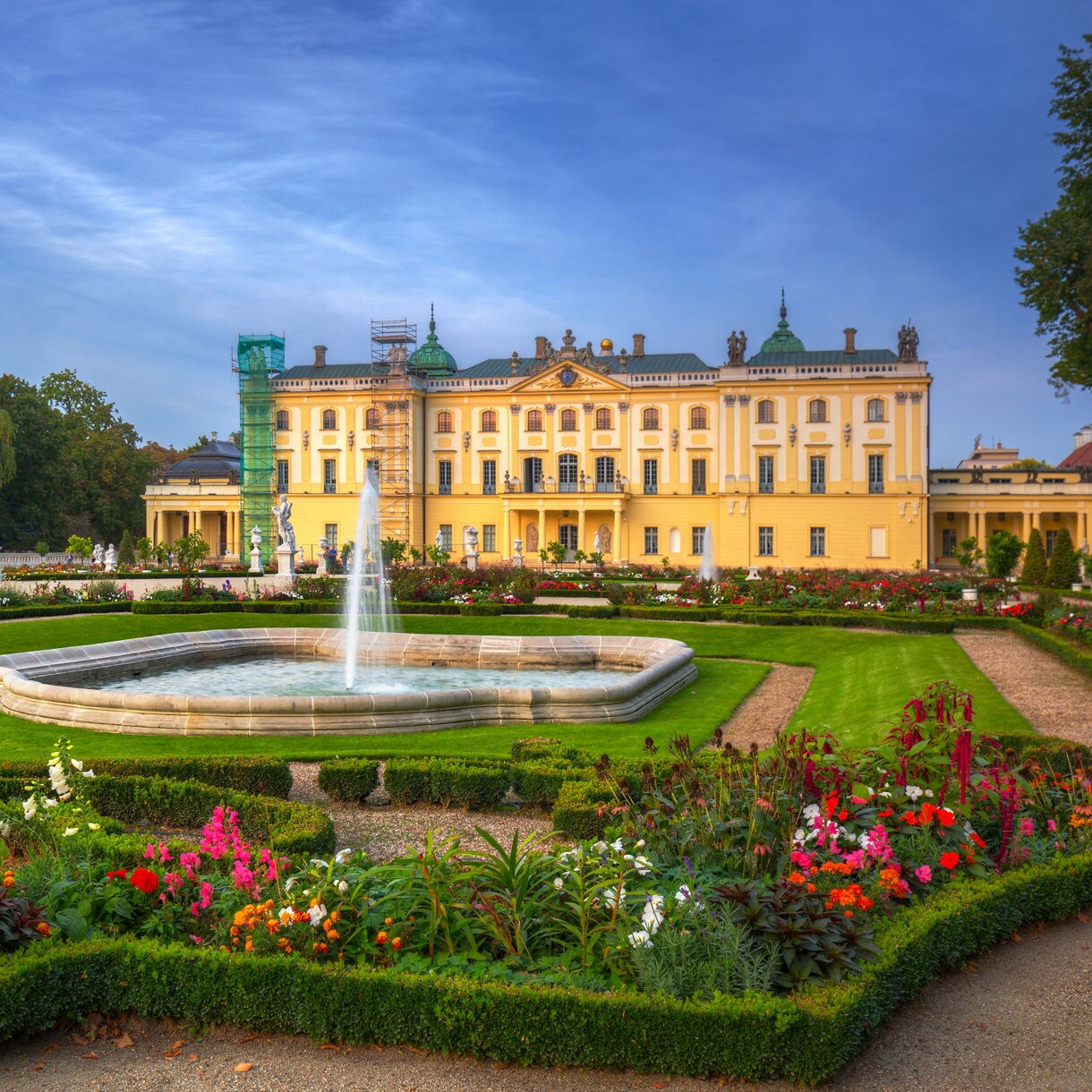Ornate yellow palace with a fountain and symmetrical French gardens filled with colorful flowers and statues under a clear blue sky.