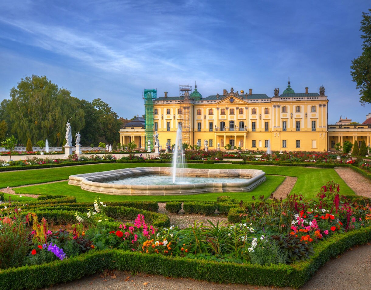 Ornate yellow palace with a fountain and symmetrical French gardens filled with colorful flowers and statues under a clear blue sky.