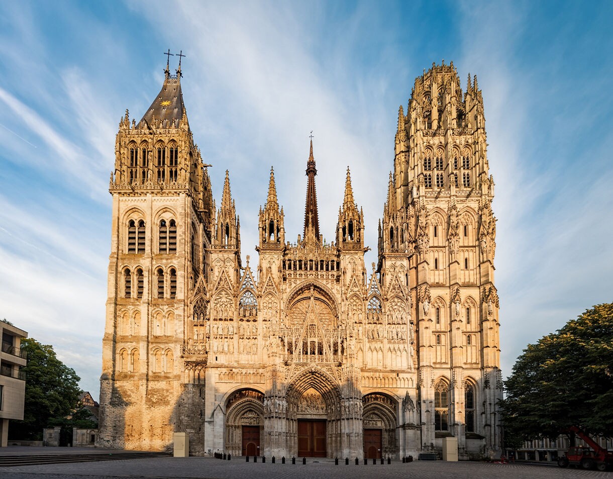Front view of Rouen Cathedral in France, showcasing ornate Gothic architecture with tall spires, elaborate carvings, and arched entrances under a bright sky.