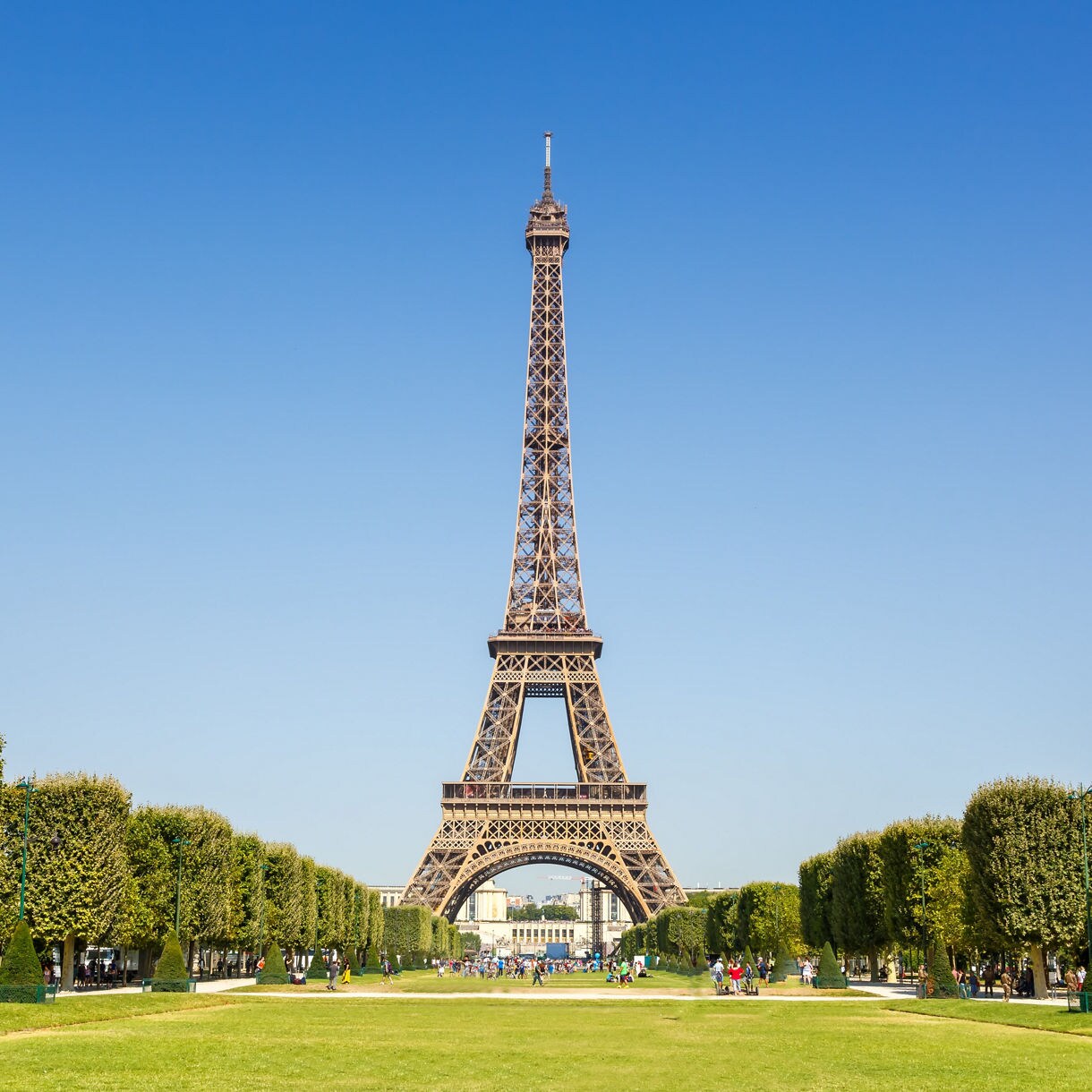 Eiffel Tower framed by manicured green lawns and tree-lined paths on a clear summer day.