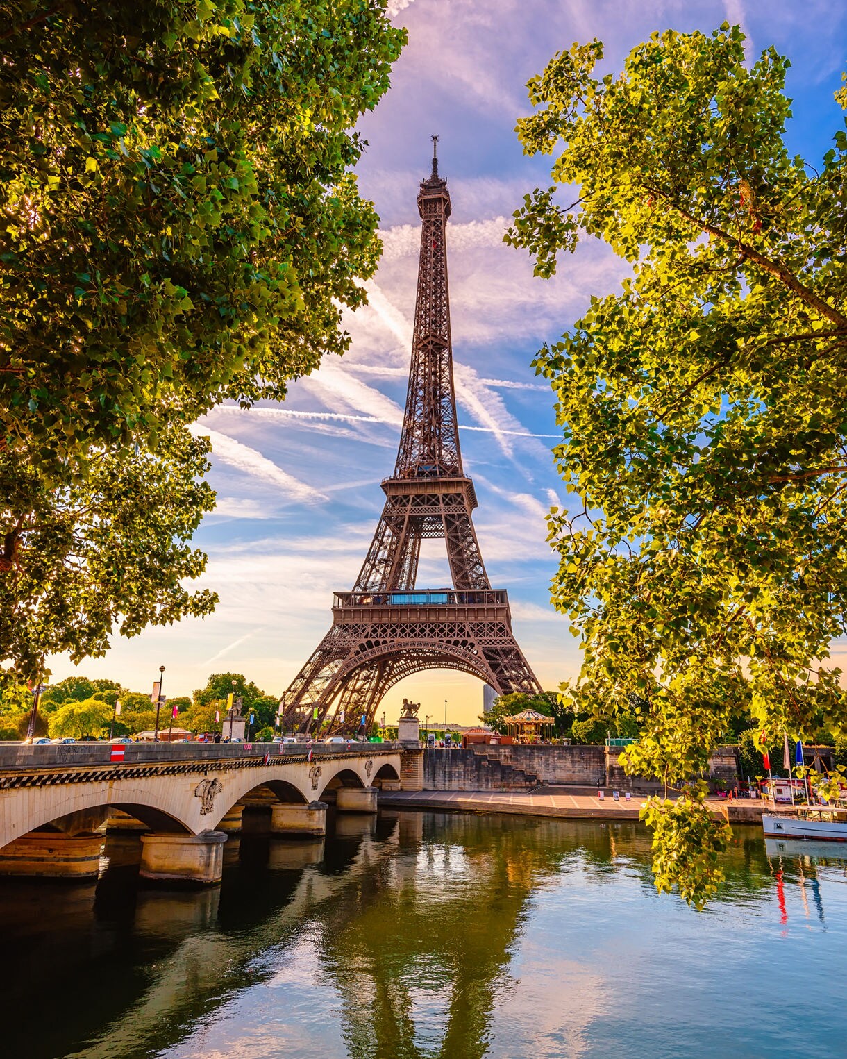 Eiffel Tower framed by leafy trees with its reflection in the Seine at sunset.