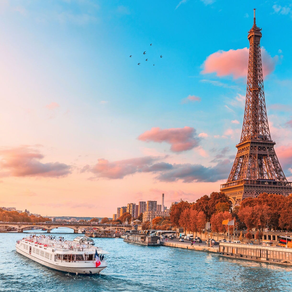 Wide view of a white cruise boat sailing along the blue waters of the Seine River in Paris with the iconic Eiffel Tower soaring over autumn trees on the left bank, under a bright sky painted with soft pink and orange clouds.