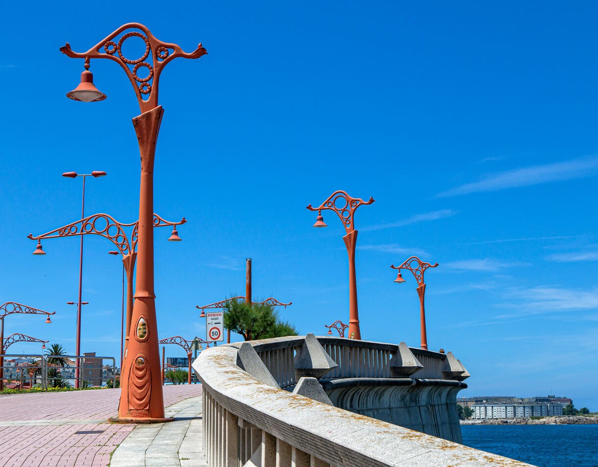 Coastal walkway in A Coruña lined with uniquely shaped orange streetlamps, curving beside the ocean under a clear blue sky.