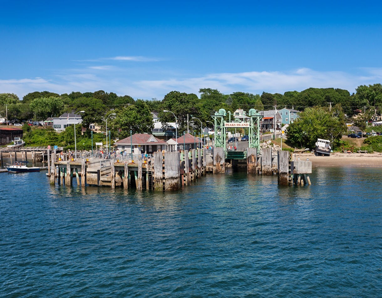 View of the Peaks Island ferry landing with a green dock structure, calm blue water and a cluster of shoreline homes framed by summer trees.