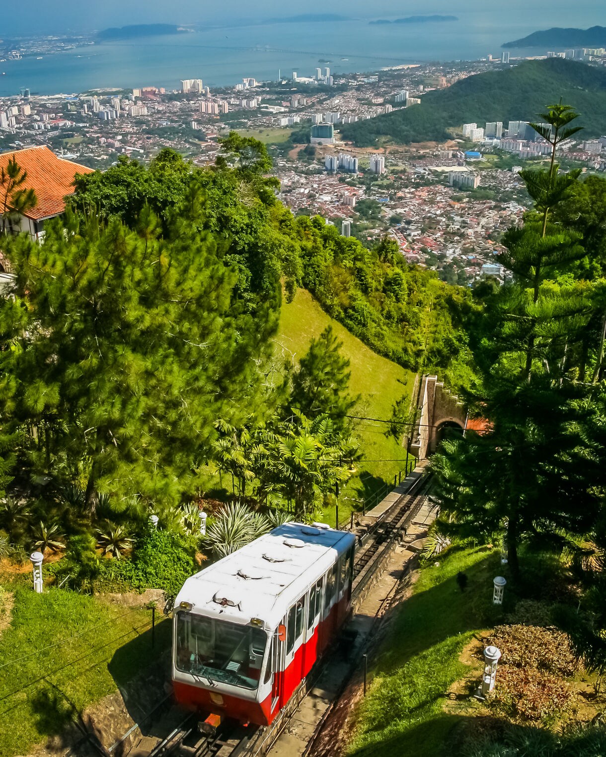 Red and white funicular train ascending Penang Hill, with lush greenery in the foreground and George Town cityscape and coastline in the distance.