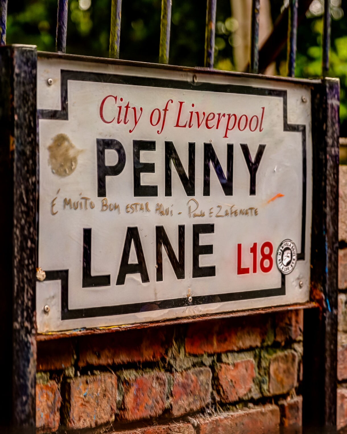Close-up of the Penny Lane street sign in Liverpool, mounted on a brick wall with iron railings and trees in the background.