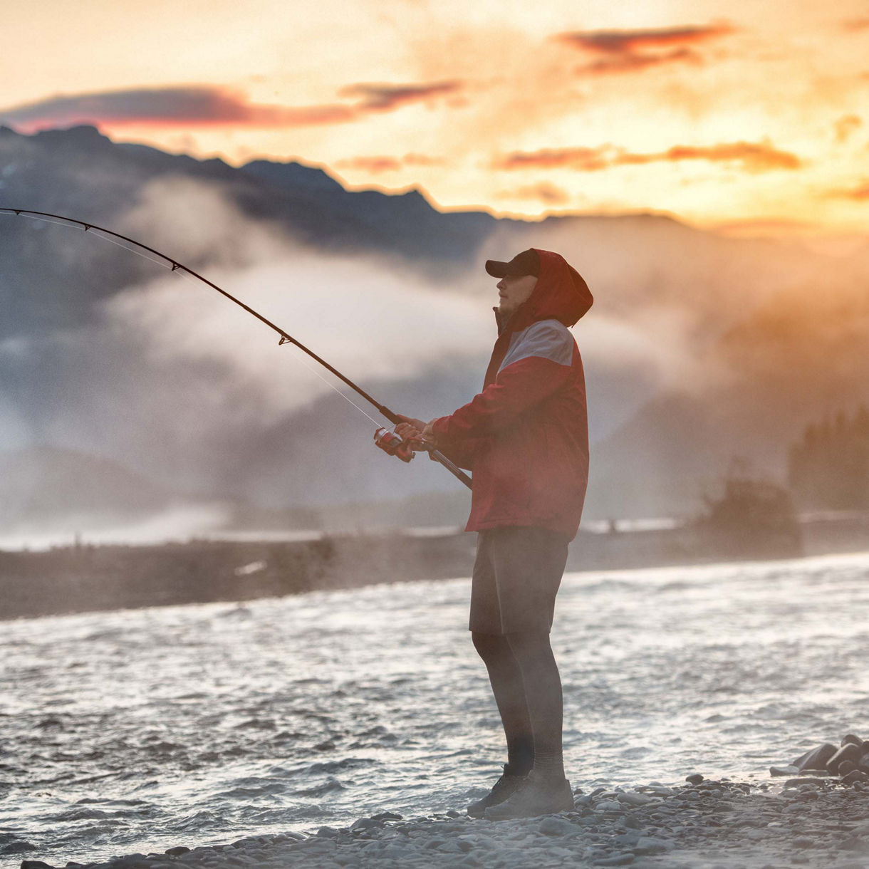 Person fishing by a river at sunset with mist rising and mountains silhouetted in the background.