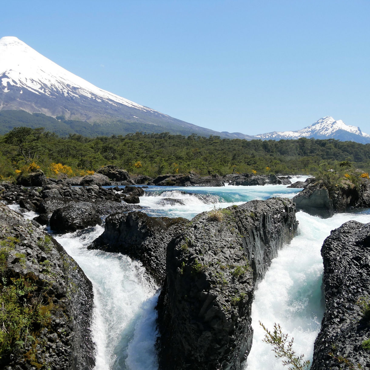 Rushing turquoise water plunging through dark volcanic rock at Petrohué Falls with the snow-covered Osorno Volcano rising in the background.