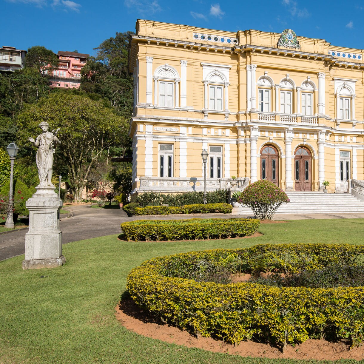 Ornate yellow neoclassical palace in Petrópolis framed by manicured gardens, hedges and a marble statue under a clear blue sky.