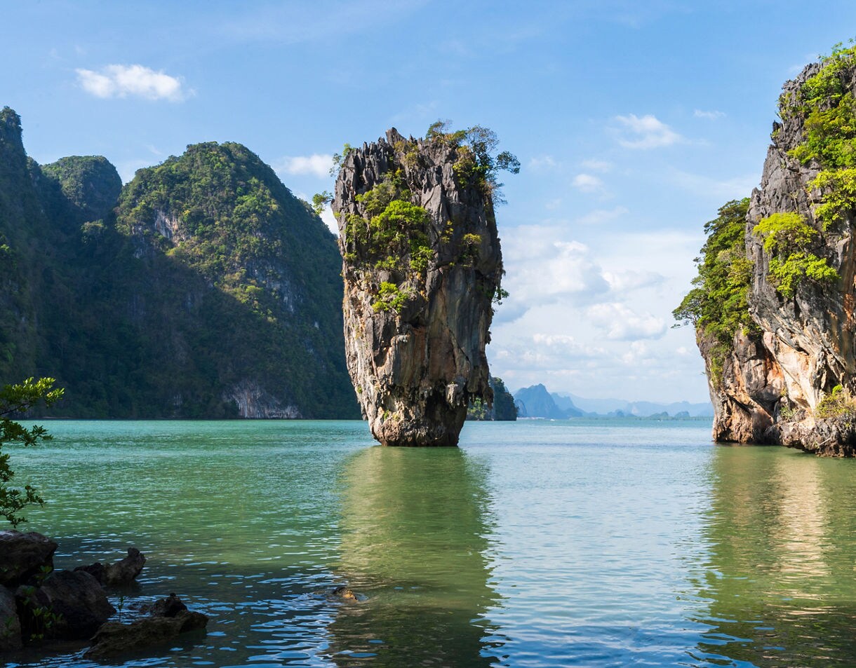 A tall, narrow limestone rock stands in calm green water, surrounded by larger forested cliffs under a bright sky.