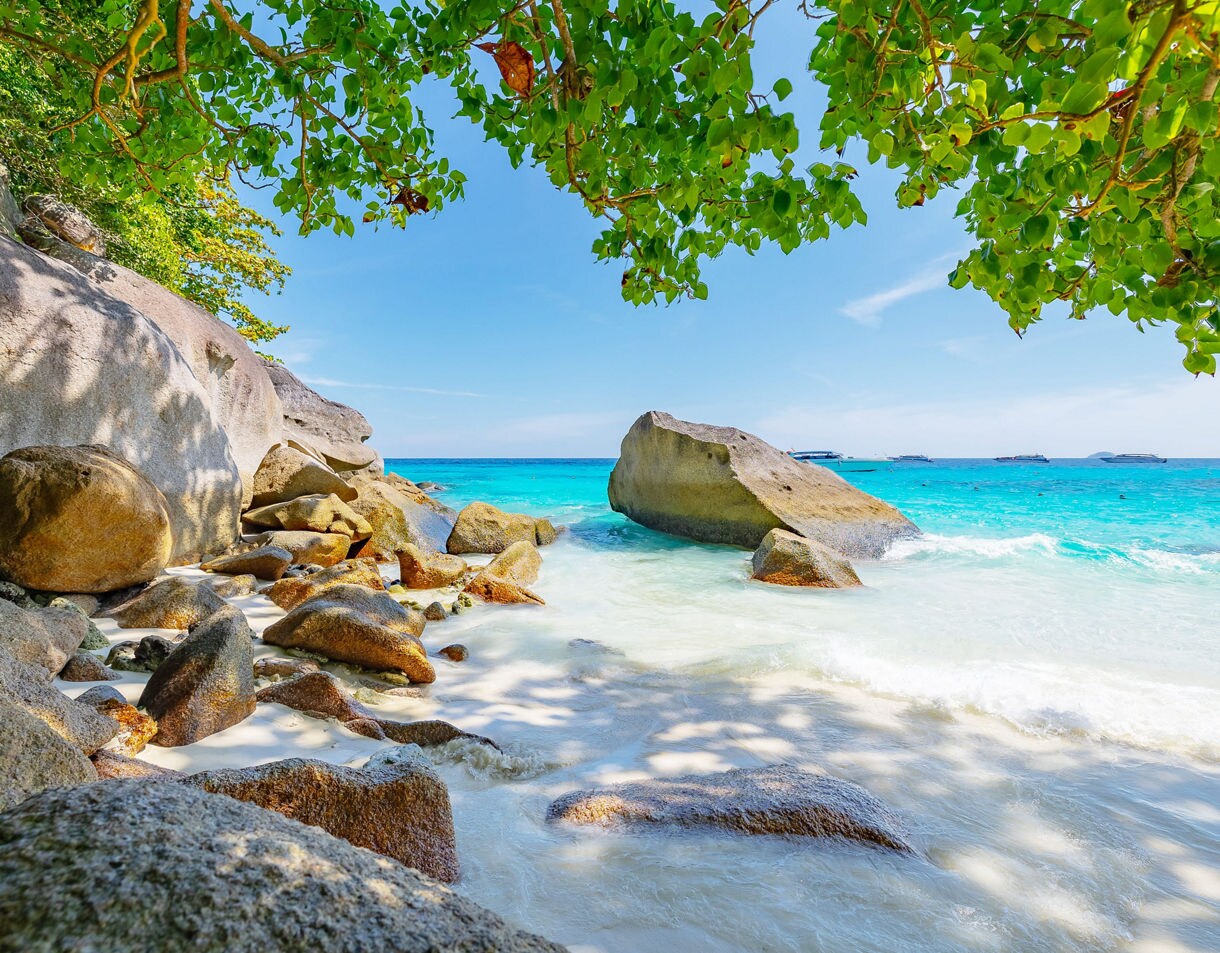 Rocky shoreline with large boulders and overhanging greenery on a bright beach in Phang Nga, facing vivid turquoise water.