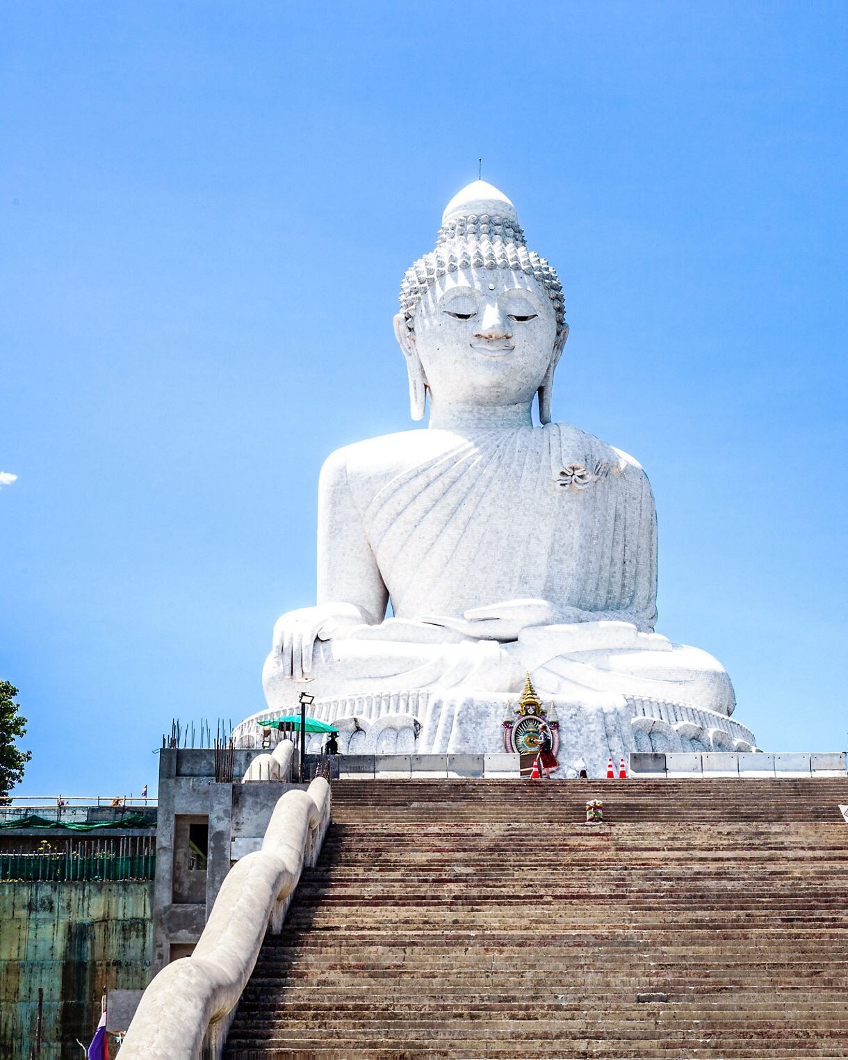 The massive white marble Big Buddha statue in Phuket viewed from the base of a wide staircase under a bright blue sky.