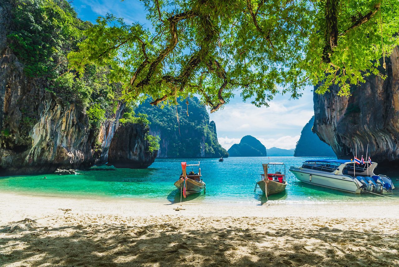 Longtail boats and a speedboat anchored on a sandy Phuket beach with emerald water and limestone cliffs in the background.