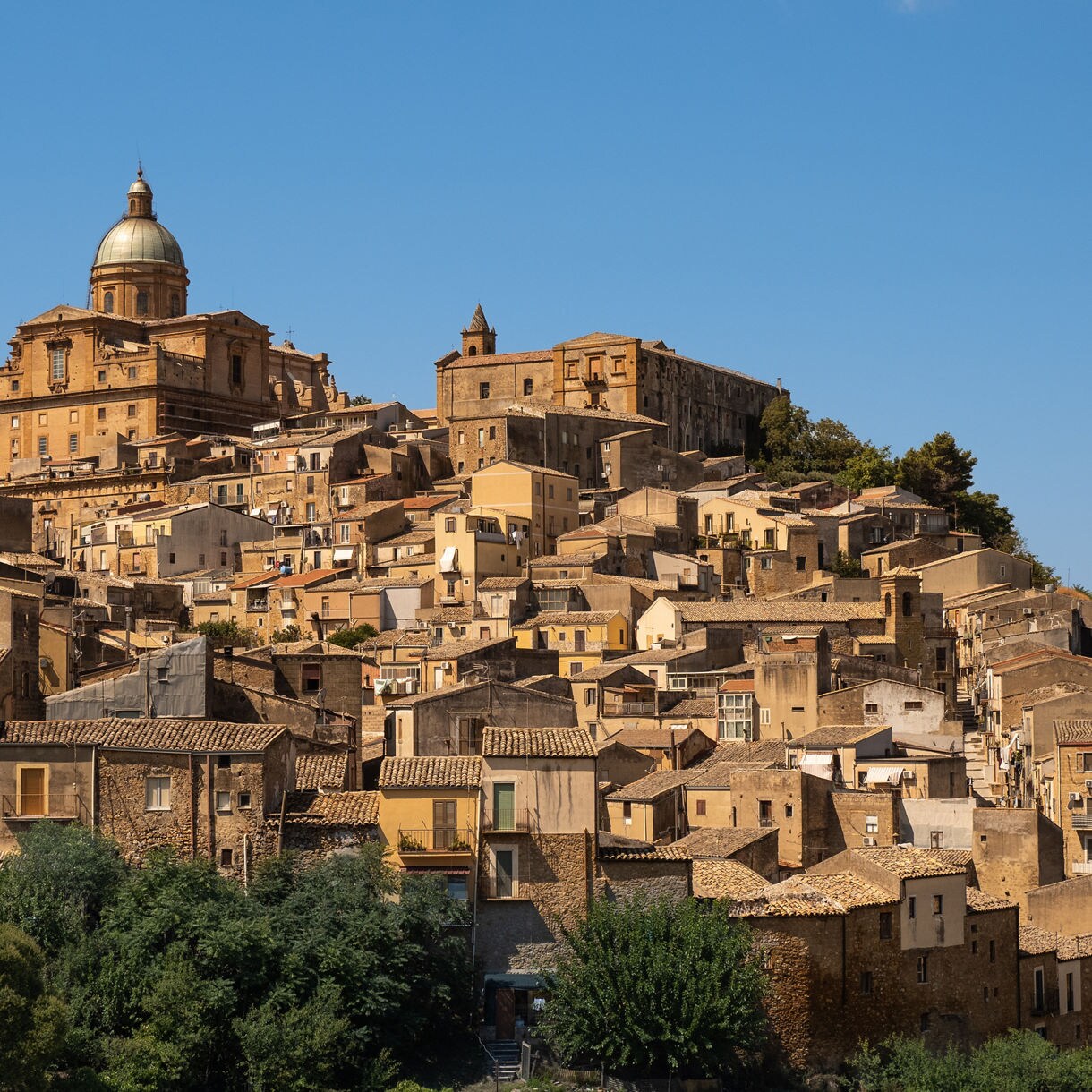 Elevated view of Piazza Armerina showing tightly packed tan stone houses stacked along a hillside, leading up to a large domed cathedral under a clear blue sky.