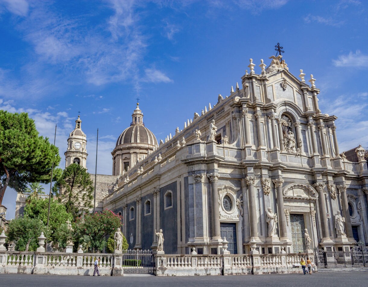 Wide view of Piazza del Duomo in Catania showing the ornate Baroque facade of the cathedral, flanked by lush trees and historic buildings beneath a bright blue sky.
