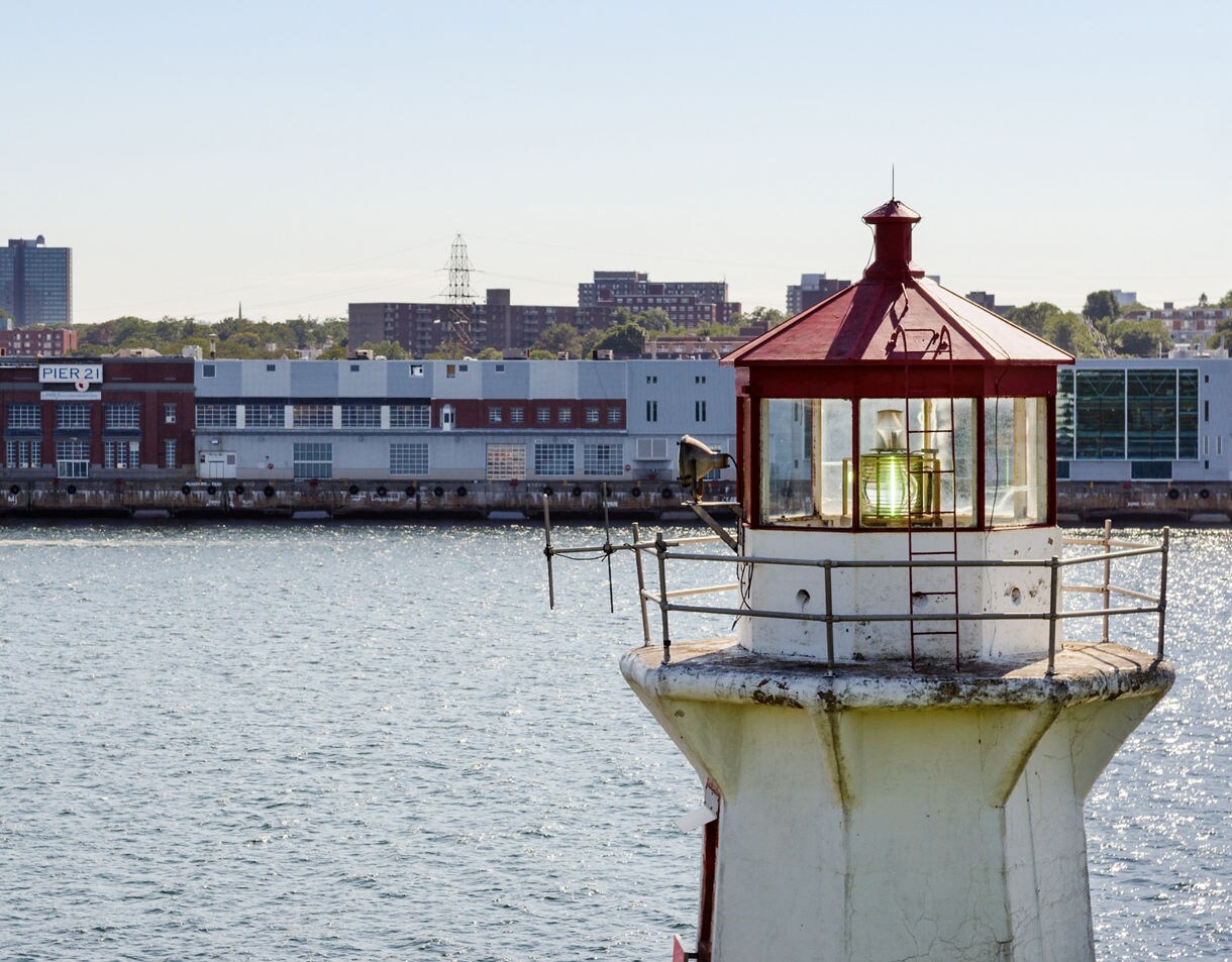 Close-up of a small white lighthouse with a red roof glowing in the sunlight, overlooking the water and Pier 21 on the Halifax waterfront.