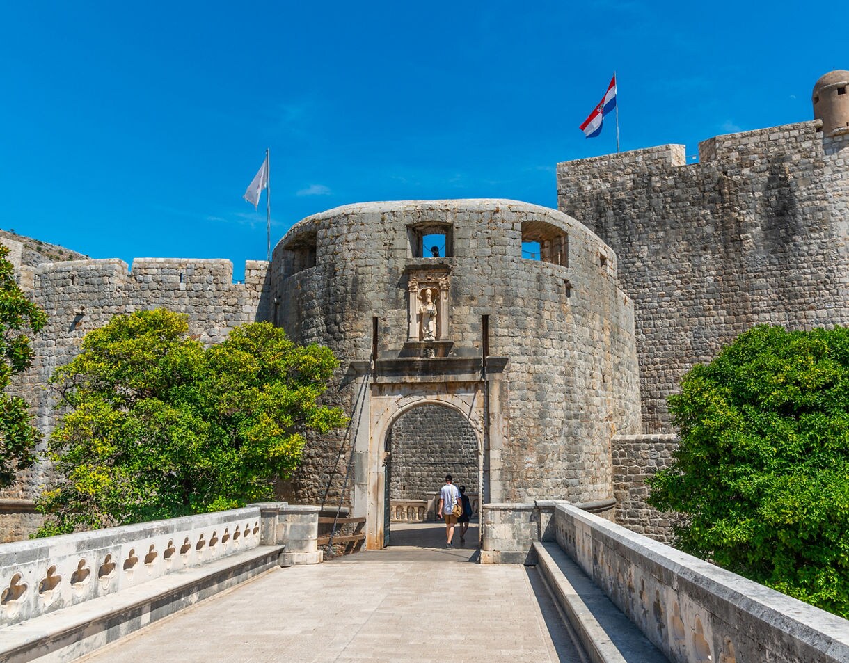 Pile Gate in Dubrovnik, Croatia, with its arched stone entrance, medieval walls and statues above the gate, framed by greenery and a bright blue sky.
