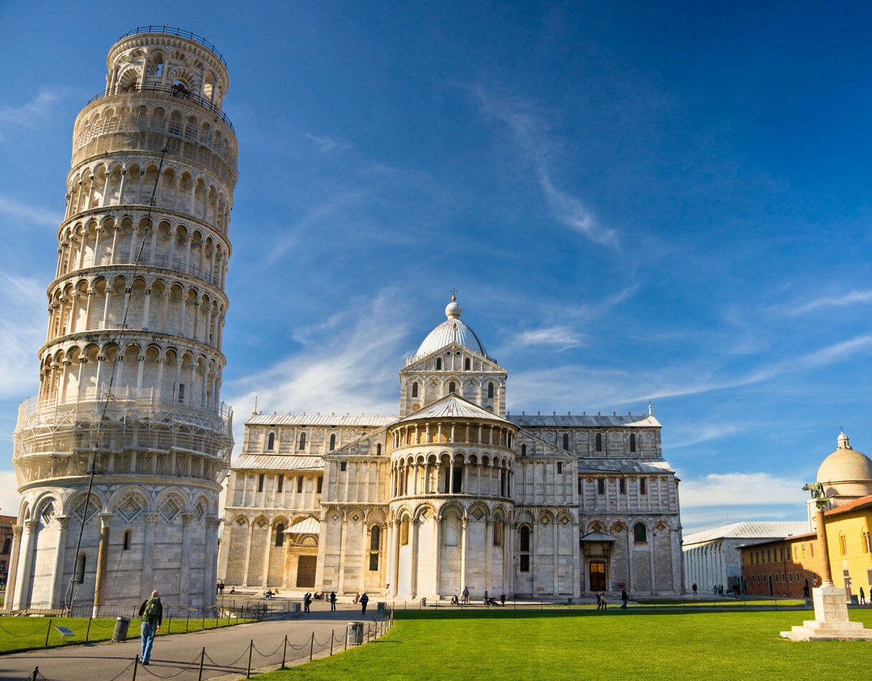  The Leaning Tower of Pisa tilted noticeably to one side next to the white marble Pisa Cathedral, with a grassy plaza and visitors walking around on a sunny day.