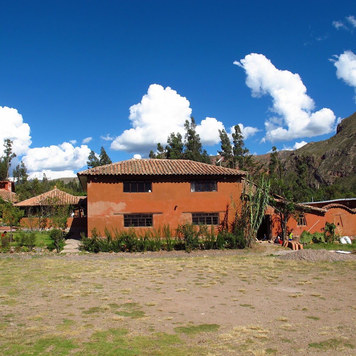 Rustic adobe buildings with terracotta roofs in a grassy courtyard, set against tall trees and mountains under a bright blue Andean sky.