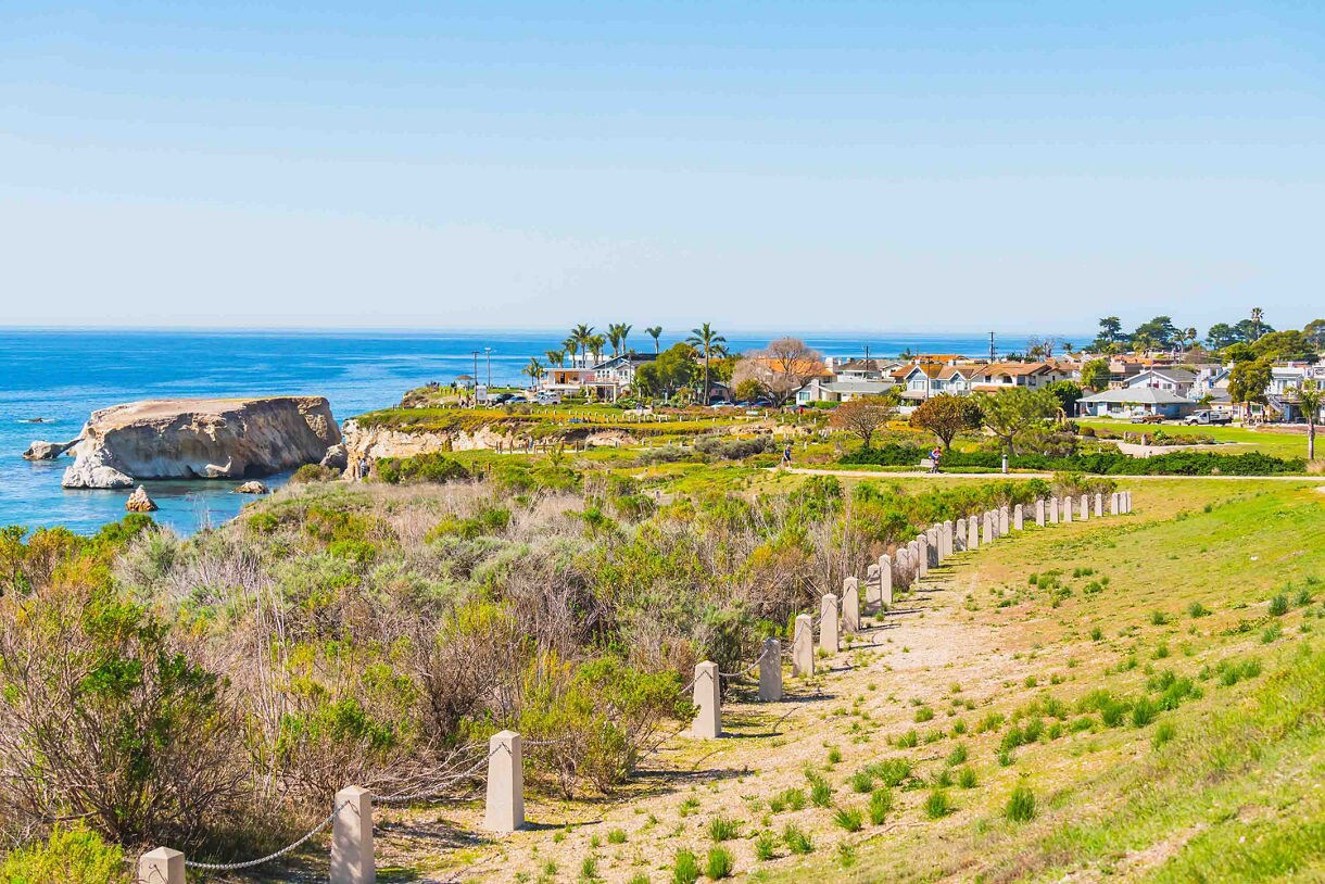 A scenic coastal view of Pismo Downtown Beach on a bright, sunny day, featuring lush green vegetation, a winding dirt path lined with wooden posts, and a rocky shoreline.
