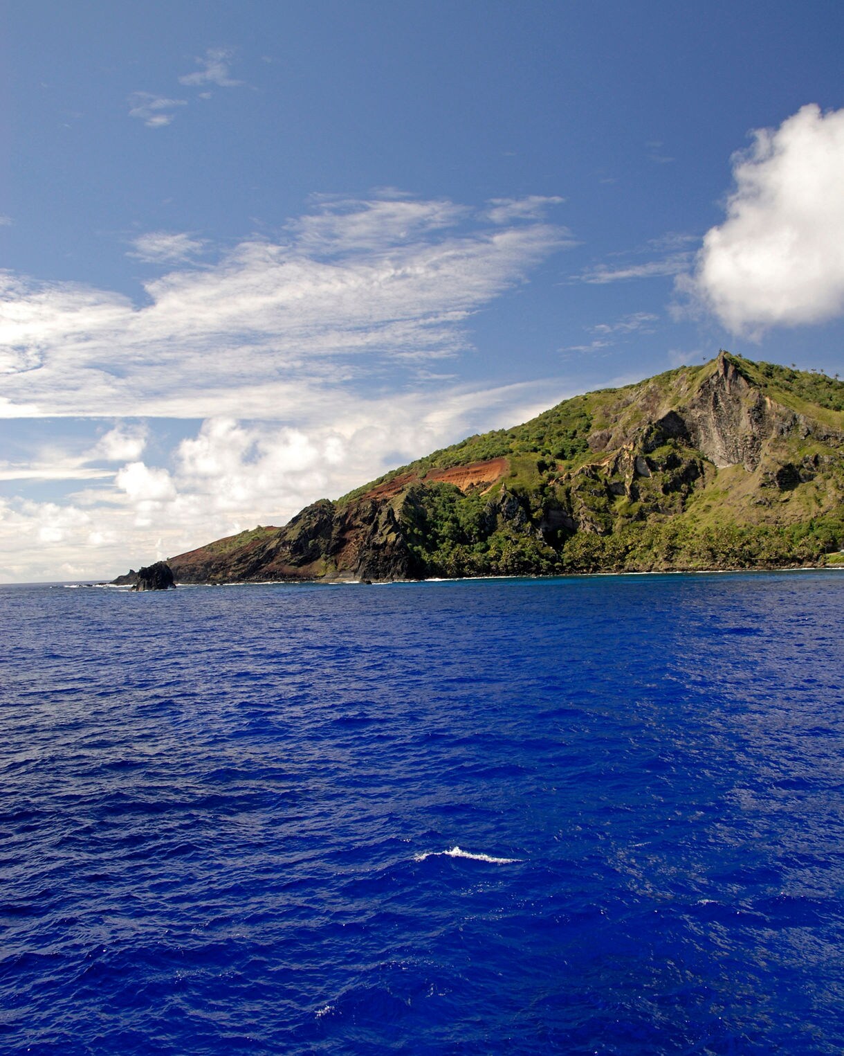 A rugged green island with steep rocky slopes rising from vivid deep blue ocean water under a bright sky with scattered clouds.