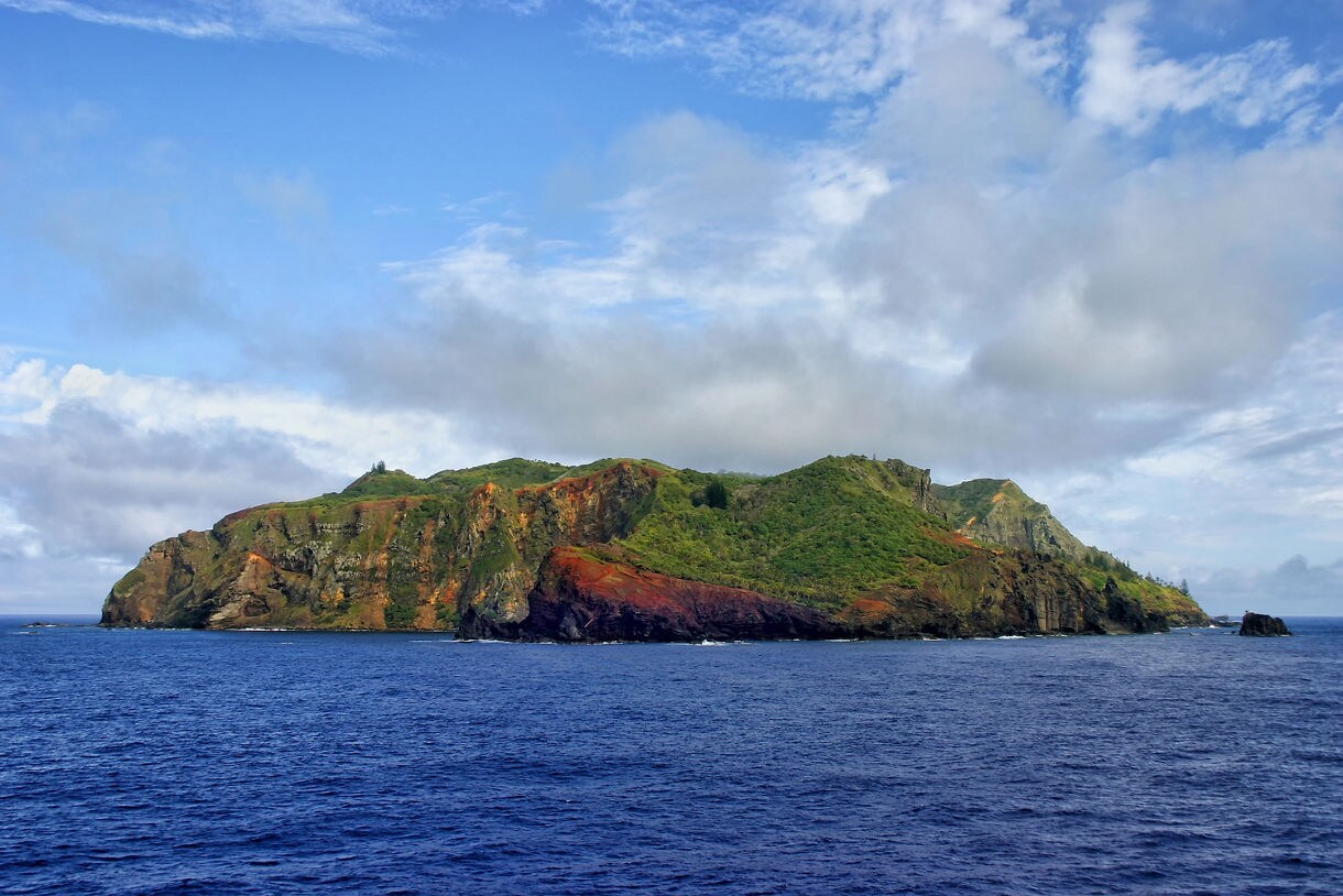 A volcanic island covered in bright green vegetation with streaks of red and brown rock, surrounded by dark blue ocean under a partly cloudy sky.