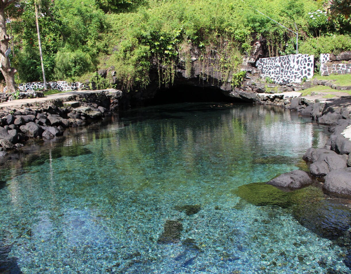 Natural freshwater pool with transparent turquoise water leading into a dark cave, surrounded by volcanic rocks and lush greenery.