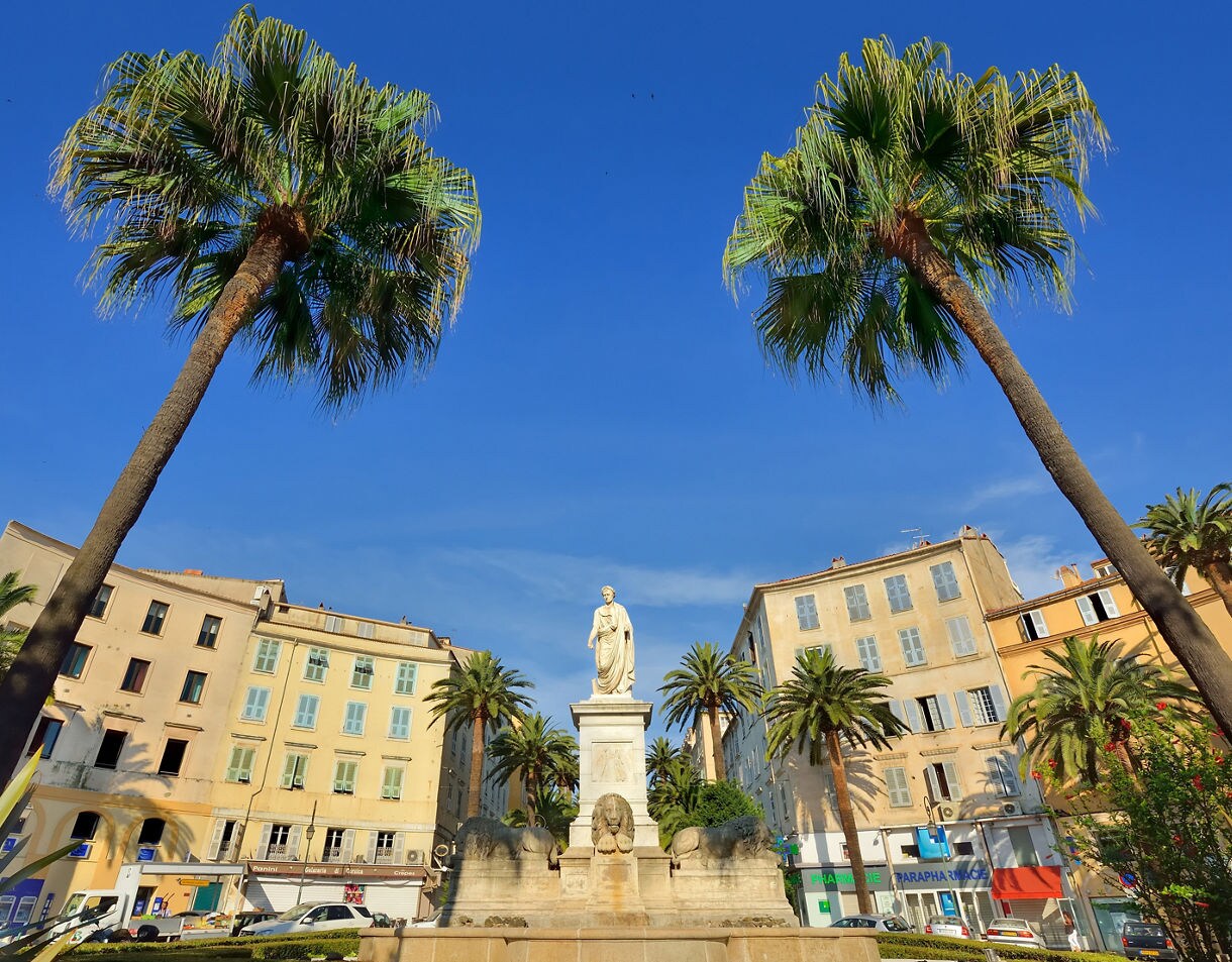 Statue of Napoleon surrounded by palm trees at Place Foch in Ajaccio, Corsica, with pastel buildings and a clear blue sky in the background.