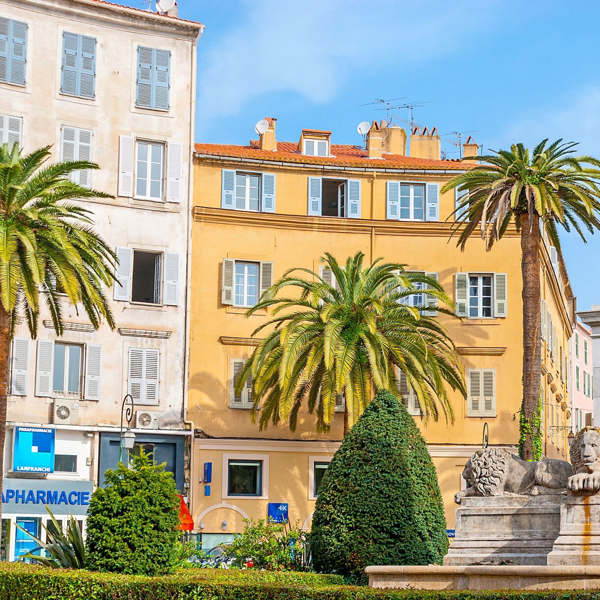 Statue of Napoleon at Place Foch in Ajaccio, France, surrounded by palm trees, lion sculptures and colorful historic buildings.