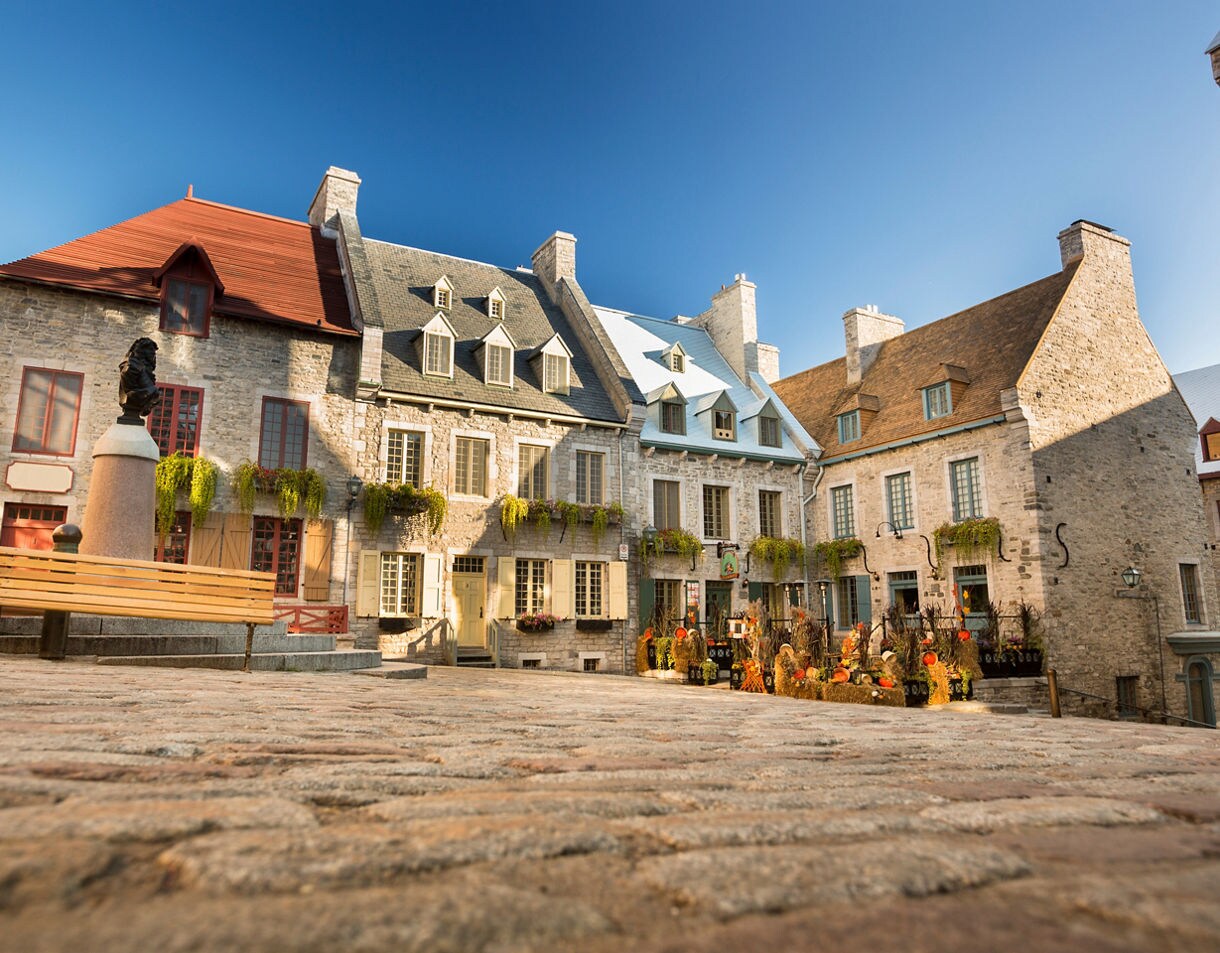 Sunlit courtyard in Place Royale with cobblestone ground, stone buildings with colorful roofs and hanging greenery.