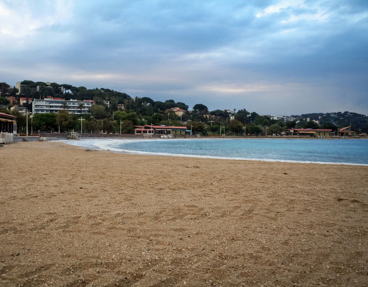 Quiet view of Plage du Mourillon featuring a wide sandy beach, calm blue water and a row of buildings and trees along the shoreline under a cloudy sky.