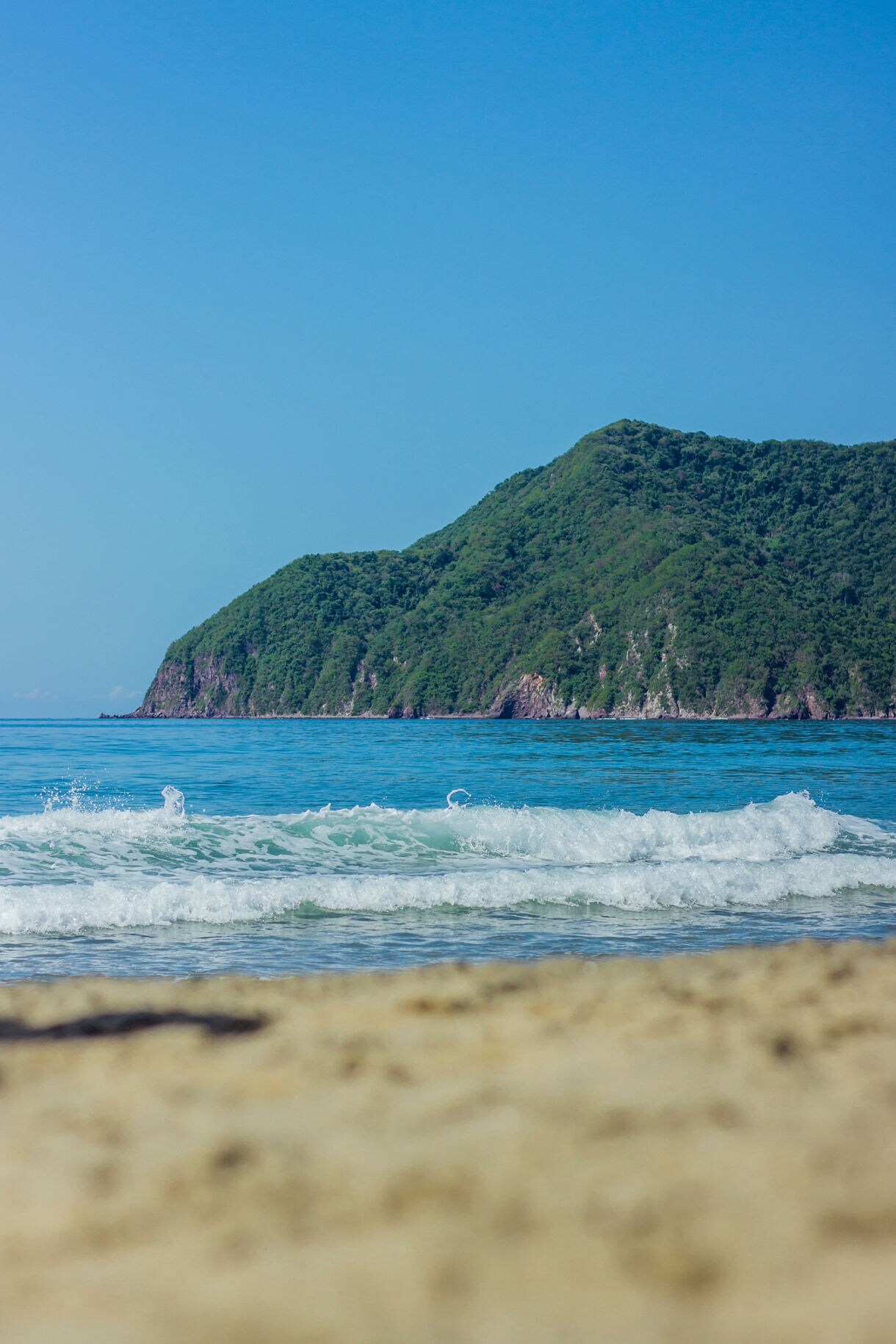 Gentle waves rolling onto sandy Playa Miramar with a backdrop of tree-covered cliffs under a clear blue sky.